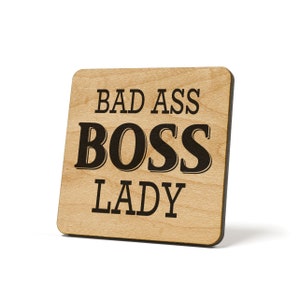 May include: Wooden sign with the text "Bad Ass Boss Lady" in black lettering.