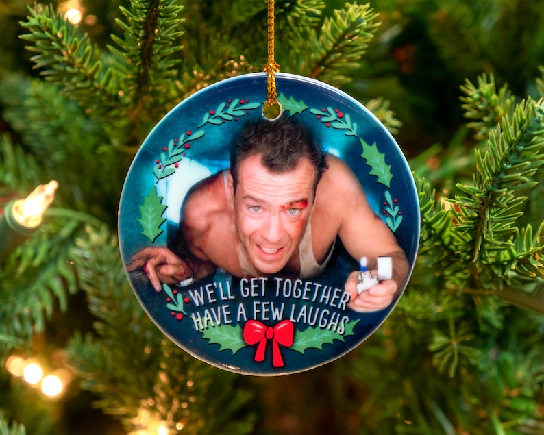 Op de afbeelding: Een ronde kerstbal met een blauwe achtergrond en een rand van een hulstkrans. De kerstbal heeft een foto van een man met een bloedneus en een aansteker in zijn hand. De tekst "We'll get together have a few laughs" staat op de kerstbal gedrukt.