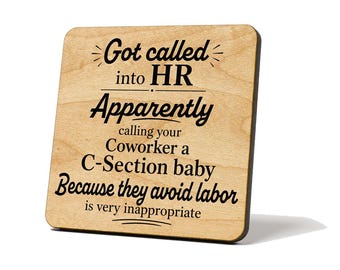 May include: A square light-colored wooden coaster with rounded corners featuring black text that reads: Got called into HR Apparently calling your Coworker a C-Section baby Because they avoid labor is very inappropriate, with decorative lines.