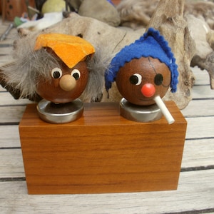 May include: Two wooden figurines with painted faces and hats. One figurine has a blue hat and a white mustache, the other has an orange hat and gray hair. Both figurines are on a wooden base.