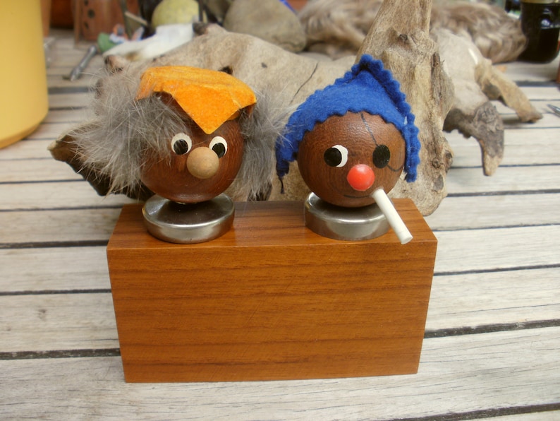 May include: Two wooden figurines with painted faces and hats. One figurine has a blue hat and a white mustache, the other has an orange hat and gray hair. Both figurines are on a wooden base.