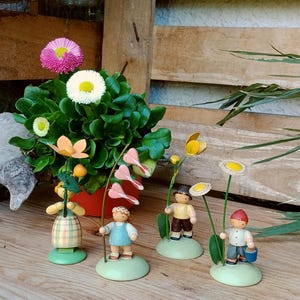 Könnte beinhalten: Eine Sammlung von Holzfiguren, die Kinder und Blumen darstellen. Die Figuren stehen auf grünen Sockeln und halten Blumenstiele. Im Hintergrund befindet sich eine Topfpflanze mit rosa und weißen Blüten. Die Szene ist auf einer Holzoberfläche aufgebaut.