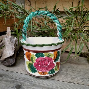 Könnte beinhalten: Weißer Keramikkorb mit geflochtenem türkisfarbenem Griff und floralem Design. Der Korb hat eine große rosa Rose, umgeben von grünen, orangefarbenen und braunen Blättern. Der Rand ist gewellt und grün eingefasst.
