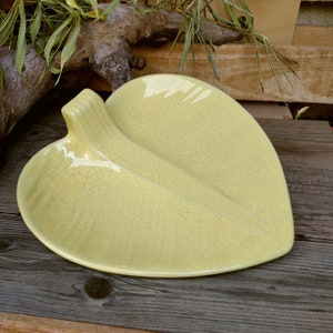 May include: A pale yellow ceramic leaf-shaped dish with a crackled glaze. The dish has a raised handle on one side and a slightly curved shape.