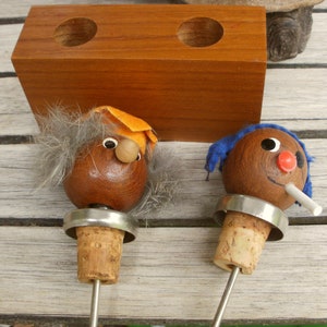 May include: Two wooden bottle stoppers with faces. One has a blue hat and a cigarette, the other has a gray beard and an orange hat. They are sitting on a wooden block with two holes.