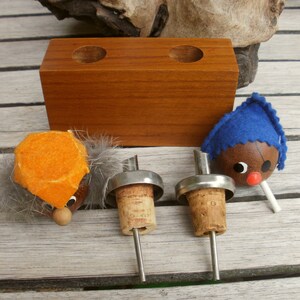 May include: Three bottle stoppers with wooden heads and felt hats. The stoppers are made of cork and metal. The heads are orange, blue, and brown. The stoppers are sitting on a wooden surface.