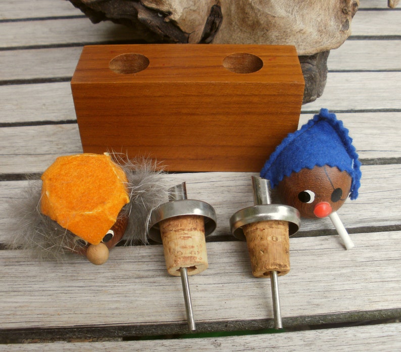May include: Three bottle stoppers with wooden heads and felt hats. The stoppers are made of cork and metal. The heads are orange, blue, and brown. The stoppers are sitting on a wooden surface.