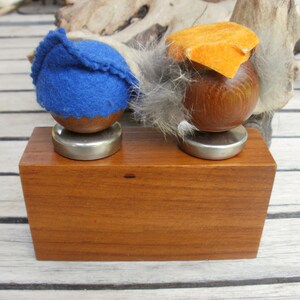 May include: Two wooden figurines with felt hats on a wooden base. The figurine on the left has a blue felt hat and the figurine on the right has an orange felt hat.