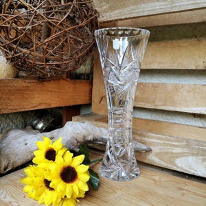 Könnte beinhalten: Eine klare, geschliffene Glasvase mit ausgestellter Oberseite und komplizierten Sternen- und geometrischen Mustern. Die Vase ist hoch und schlank, ideal für die Präsentation von Blumen. Helle gelbe Sonnenblumen mit braunen Zentren liegen in der Nähe der Vase.