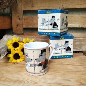 Puede incluir: Una taza de cerámica beige con un diseño de ratón de dibujos animados, un sombrero de copa negro y una pajarita roja. La taza está junto a dos cajas con el mismo diseño. Girasoles amarillos en el fondo.