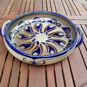 May include: A round ceramic serving dish with a blue rim and handles. The interior features a brown and blue floral pattern on a beige background. The dish is sitting on a wooden table.