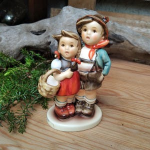 May include: A ceramic figurine of two children, a boy and a girl, standing close together. The girl wears a red dress and holds a basket. The boy wears a hat, a blue jacket, and a red scarf. The figurine is on a round, off-white base.