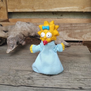 Puede incluir: Una figura amarilla de Maggie Simpson con un vestido azul claro y un lazo azul. La figura tiene un chupete rojo en la boca y posa con los brazos extendidos. Es un artículo de colección.