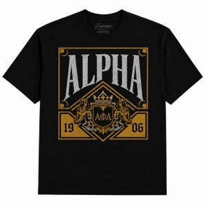 May include: A black short-sleeve t-shirt featuring a large graphic print. The design includes the word ALPHA in silver, a gold crest with two sphinxes, a crown, the Greek letters ΑΦΑ, and the numbers 19 and 06 centered within a gold diamond frame.