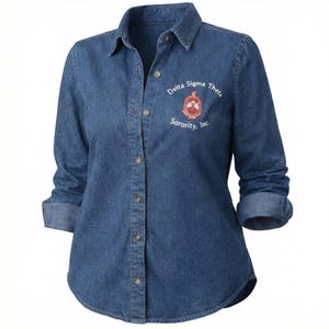 May include: A long-sleeved, button-down denim shirt in a medium blue wash. The shirt features rolled-up sleeves and the embroidered text "Delta Sigma Theta Sorority, Inc." in white and red on the left chest.