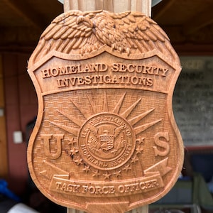 May include: A wooden badge with the words "Homeland Security Investigations" and "Task Force Officer" carved into it. The badge also features a carved eagle and the U.S. Department of Homeland Security seal.