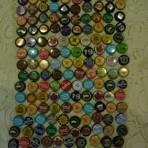 May include: A display of assorted bottle caps, each with unique branding and designs. The caps are tightly packed, displaying a range of colours like red, blue, green, and gold. Several caps feature text, including "19", "Angry Orchard", and "Corona Extra".