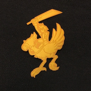 May include: A gold-coloured embroidered patch featuring a stylised image of a knight riding a bird-like creature, holding a sword.