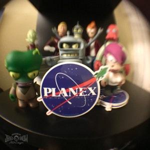 Puede incluir: Primer plano de una pegatina azul y roja con la palabra "PLANEX" en letras blancas. La pegatina est&aacute; rodeada de otras pegatinas y peque&ntilde;as figuritas, incluyendo un extraterrestre verde, un robot y un personaje de dibujos animados.
