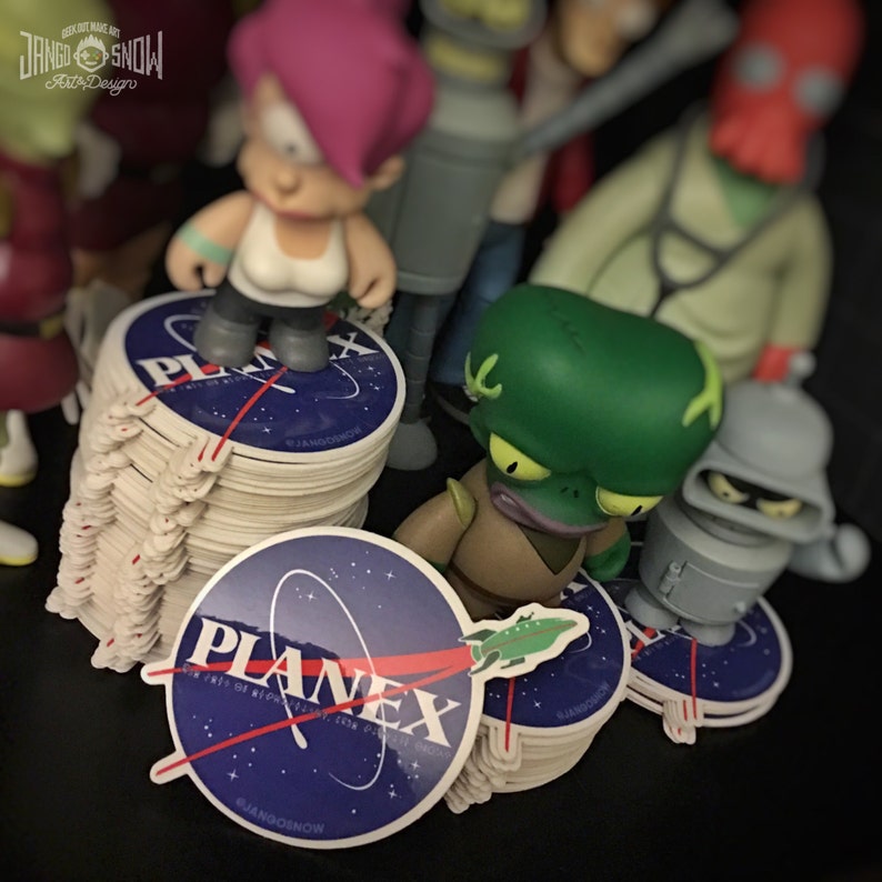 Puede incluir: Una pila de pegatinas redondas con un dise&ntilde;o azul, blanco y rojo que presenta el texto "PLANEX" y un cohete. Las pegatinas est&aacute;n sobre una superficie negra con varias figuritas peque&ntilde;as.