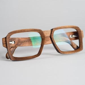 Puede incluir: Un par de gafas con montura de madera marrón y lentes transparentes. Las monturas son rectangulares y tienen un borde ligeramente redondeado. Las palabras "HANDMADE IN" están grabadas en la montura izquierda.