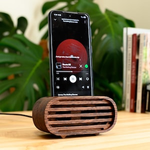 Könnte beinhalten: Ein dunkelbrauner Smartphone-Ständer aus Holz mit horizontalem Lamellendesign an der Vorderseite. Der Ständer verfügt über einen Schlitz an der Oberseite, um ein Smartphone vertikal zu halten, das eine Musik-App mit rotem Album-Cover anzeigt.