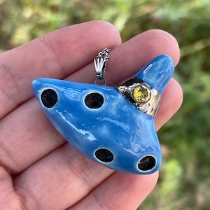 Könnte beinhalten: Blauer Keramik-Okarina-Anhänger mit silberfarbenem Metallakzent und gelbem Edelstein. Der Anhänger hat vier schwarze Löcher und hängt an einer silbernen Kette. Die Okarina ist ein Musikinstrument.