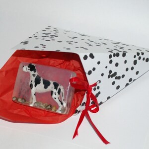 Puede incluir: Una figurita de perro d&aacute;lmata blanco y negro dentro de una bolsa de regalo blanca con cinta roja y papel de seda rojo.