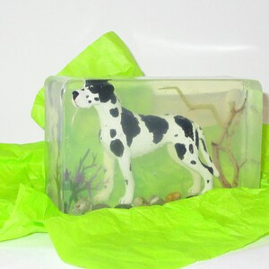 Puede incluir: Un bloque de resina transparente con una figurita de Gran Dan&eacute;s en blanco y negro en su interior. La figurita est&aacute; de pie sobre una peque&ntilde;a rama y hay plantas verdes y marrones en la resina. El bloque est&aacute; sobre un papel de seda verde.