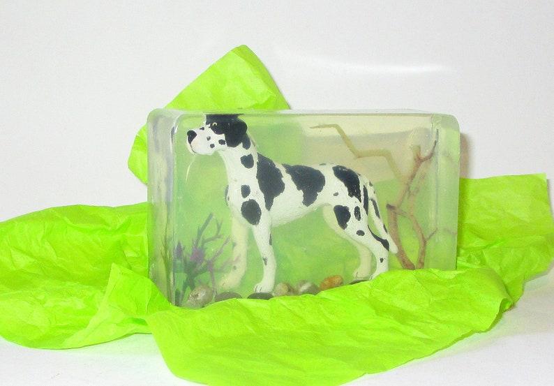 Puede incluir: Un bloque de resina transparente con una figurita de Gran Dan&eacute;s en blanco y negro en su interior. La figurita est&aacute; de pie sobre una peque&ntilde;a rama y hay plantas verdes y marrones en la resina. El bloque est&aacute; sobre un papel de seda verde.