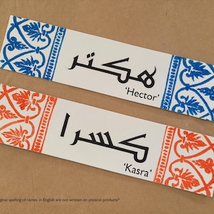 May include: Two rectangular name tags with Arabic script and English transliterations. One tag has blue floral border, the other has an orange floral border. The names "Hector" and "Kasra" are printed on the tags.