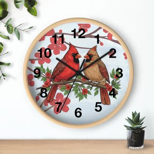 Könnte beinhalten: Eine runde Wanduhr mit hellem Holzrahmen. Das Zifferblatt zeigt zwei Kardinäle auf einem Ast mit roten Blüten. Schwarze Zahlen markieren die Stunden vor einem hellblauen Hintergrund.
