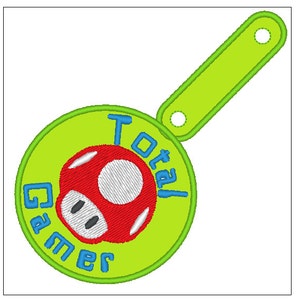 Puede incluir: Un parche bordado verde y azul con un hongo rojo y el texto "Total Gamer". El parche tiene un asa verde con dos agujeros.