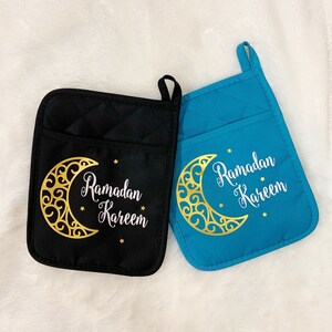 May include: Two quilted pot holders, one black and one teal, with a gold crescent moon design and the words "Ramadan Kareem" in white. Each pot holder has a pocket and a loop for hanging.