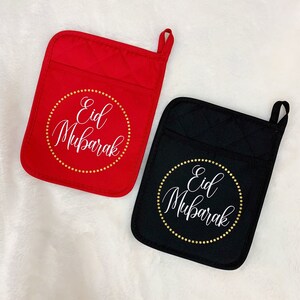 May include: Two pot holders with a quilted design. One is red and the other is black. Both have a white circle with gold stars around it and the text "Eid Mubarak" in white.
