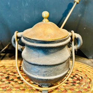 May include: A matte black cast iron cauldron with a brass lid and a matching brass handle. The pot features three short legs and two side handles. A long, thin brass stirring rod with a spherical finial rests inside the pot.