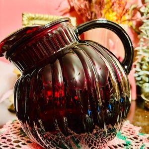 May include: A dark red glass pitcher with a ribbed design. The pitcher has a black handle and a spout for pouring. The pitcher is sitting on a pink and green doily. The pitcher is a decorative item.