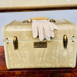 May include: A rectangular cream-colored hard-shell vanity case with a marbled texture, featuring a top handle, brass-toned metal latches, and a central lock plate. A pair of pale pink fabric gloves rests on top of the handle and the case lid.