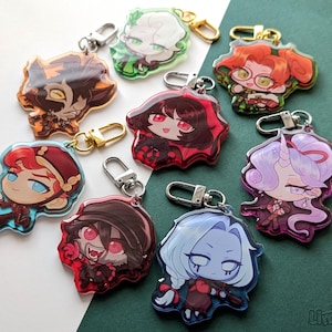 May include: A collection of colorful acrylic keychains featuring anime-style character designs. Each keychain has a unique character with different hair colors and outfits, attached to a metal clasp. The keychains are on a green surface.