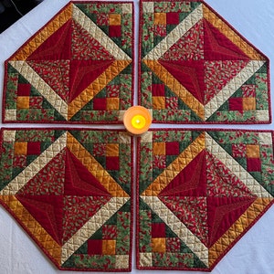 May include: Four red, green, and gold quilted placemats with a star design in the center. The placemats are arranged in a square shape around a candle.