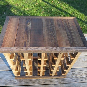 Puede incluir: Un estante de vino de madera r&uacute;stico con una superficie superior que se puede utilizar como mesa. El estante est&aacute; hecho de madera recuperada y tiene un acabado natural. La superficie superior est&aacute; hecha de varias tablas de madera que est&aacute;n unidas. El estante tiene una forma rectangular y mide aproximadamente 61 cm de ancho y 46 cm de profundidad.