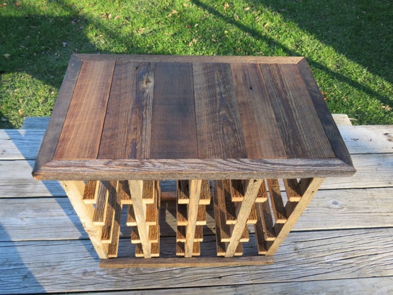 Puede incluir: Un estante de vino de madera r&uacute;stico con una superficie superior que se puede utilizar como mesa. El estante est&aacute; hecho de madera recuperada y tiene un acabado natural. La superficie superior est&aacute; hecha de varias tablas de madera que est&aacute;n unidas. El estante tiene una forma rectangular y mide aproximadamente 61 cm de ancho y 46 cm de profundidad.