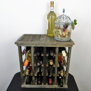 Puede incluir: Un estante de vino de madera r&uacute;stica con 18 ranuras para botellas. El estante est&aacute; te&ntilde;ido de un color marr&oacute;n oscuro y tiene un acabado desgastado. Una botella de vino blanco y una jaula de p&aacute;jaros decorativa llena de corchos de vino est&aacute;n encima del estante.
