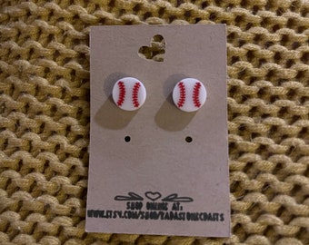 May include: A pair of white baseball stud earrings with red stitching. The earrings are displayed on a brown card with the text "Shop Online At: www.etsy.com/shop/YarnasOneCrafts".
