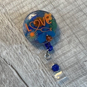 Puede incluir: Un portagafete de resina azul y naranja con un estetoscopio y la palabra "LOVE" en letras naranjas. El portagafete tiene un carrete retráctil azul y plateado y un amuleto azul con purpurina.