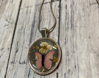 Puede incluir: Un collar de cadena de plata con un colgante redondo que presenta una mariposa rosa y flores blancas prensadas en resina transparente. El colgante tiene un fondo brillante.