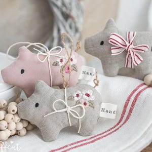 Pode incluir: Três enfeites de porco feitos à mão. Um é rosa e dois são cinzentos. Cada porco tem um desenho floral costurado e uma pequena etiqueta que diz "Hand". Um porco cinzento tem um laço listrado vermelho e branco e um coração preto.