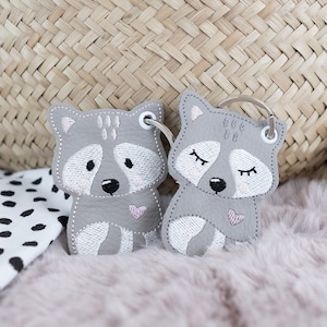 May include: Two grey raccoon-shaped keychains with pink hearts and embroidered details. The keychains are on a fluffy pink surface.