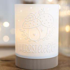 Peut inclure: Un photophore cylindrique blanc avec un motif de champignons et le texte "Herbstlicht". La base est de couleur marron clair avec des lignes verticales. Le photophore est posé sur une surface en bois.