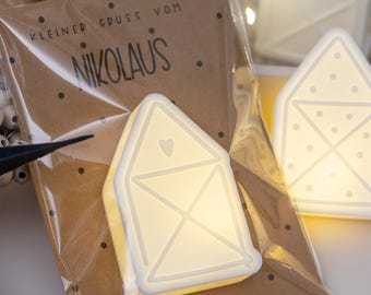 Puede incluir: Luces LED blancas en forma de casa con diseño de corazón, empaquetadas en plástico transparente y papel marrón. El texto "Kleiner Gruss vom Nikolaus" está impreso en el papel. Una estrella negra y cuentas blancas en el fondo.